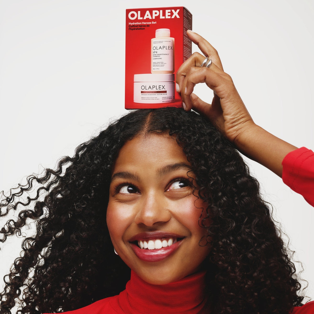 Smiling woman with curly hair balancing the Olaplex Hydration Heroes gift set on her head