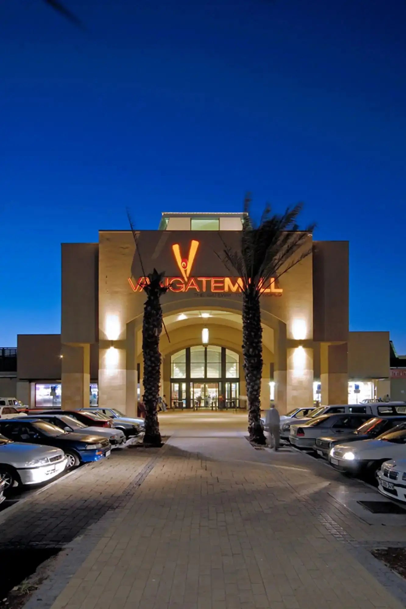 Illuminated entrance to a shopping mall with ’V’ signage and palm trees flanking the doorway.