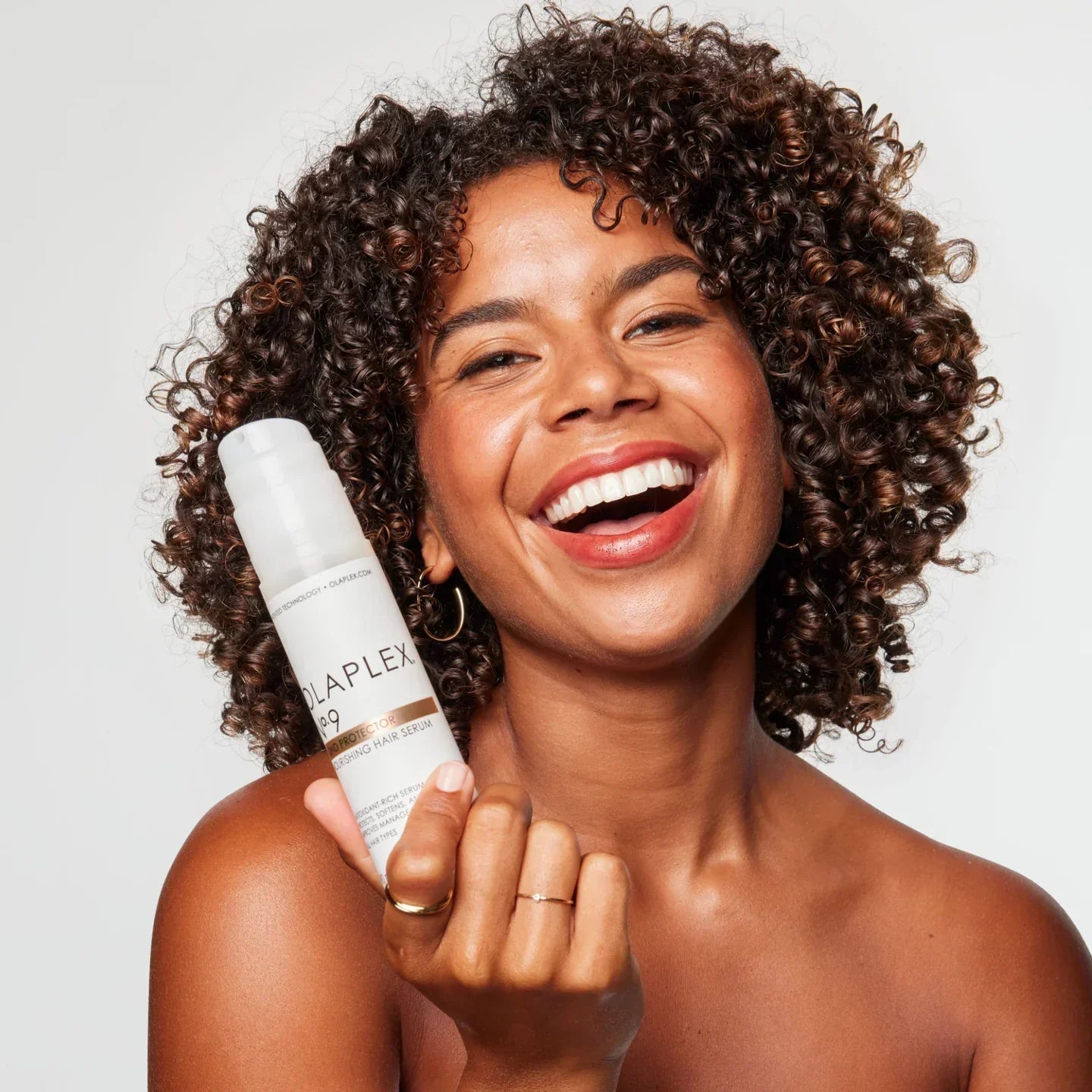 Happy woman with curly hair showcasing Olaplex No. 9 Bond Protector serum for frizz control & healthy curls.