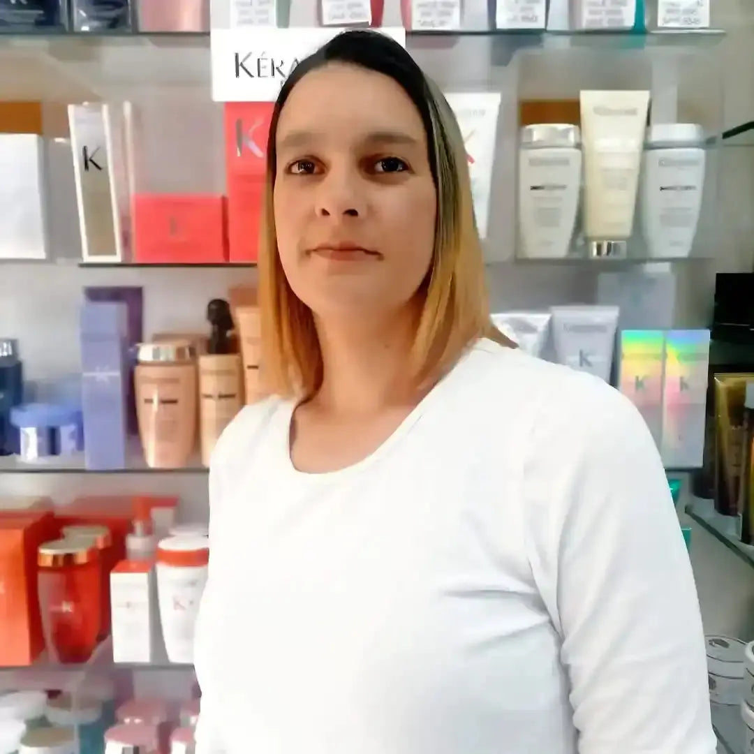 A person with blonde ombre hair wearing a white top standing in front of beauty product shelves.
