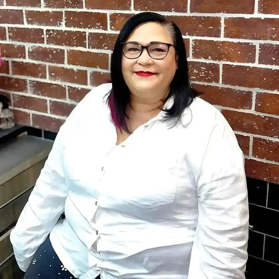 A smiling person wearing glasses and a white button-up shirt against a brick wall.