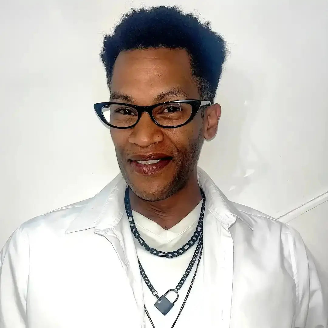 A person wearing glasses, a white shirt, and layered chain necklaces smiles at the camera.
