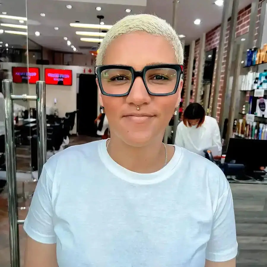 A person with platinum blonde short hair wearing black-rimmed glasses and a white t-shirt.