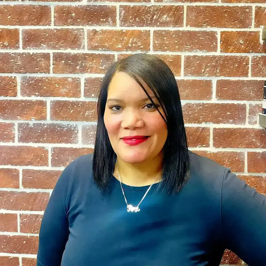 A person with straight black hair wearing a navy blue top and silver necklace against a brick wall.