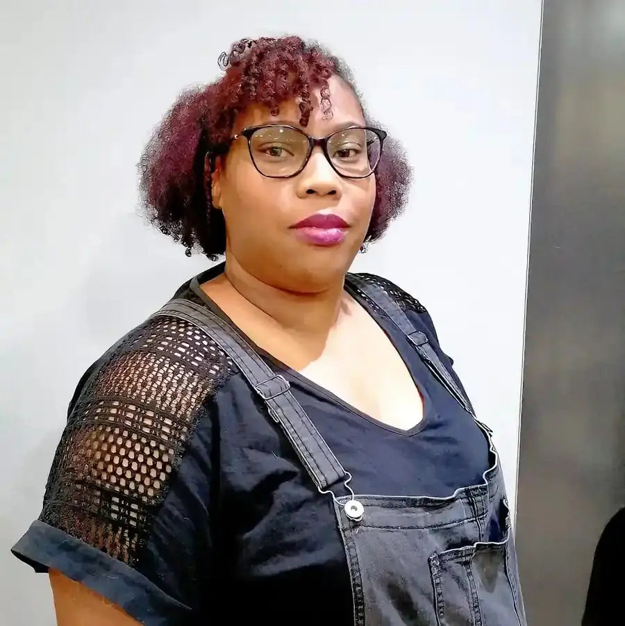 Portrait photo showing someone wearing glasses, burgundy-colored curly hair, and denim overalls with a black mesh-sleeved top.