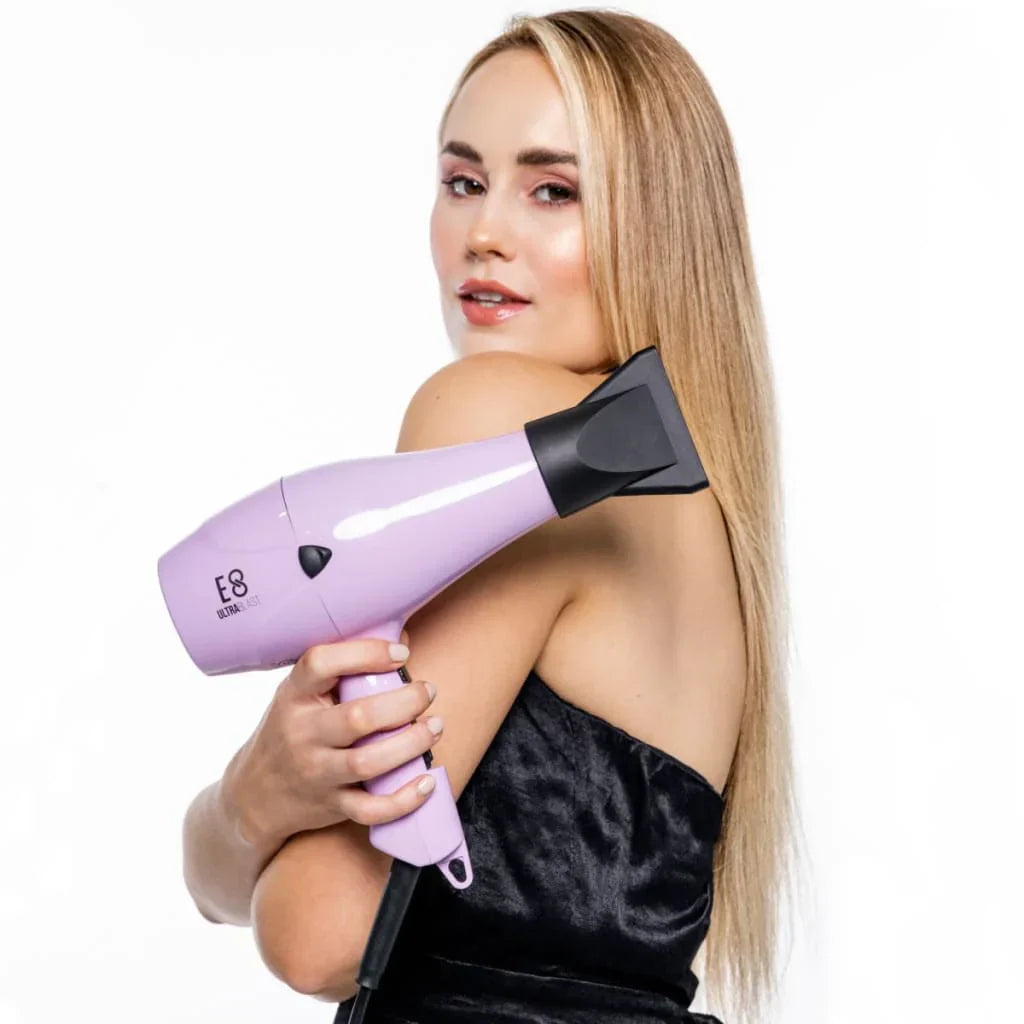 Woman with long blonde hair holds a purple hair dryer next to a flat iron and curling iron.