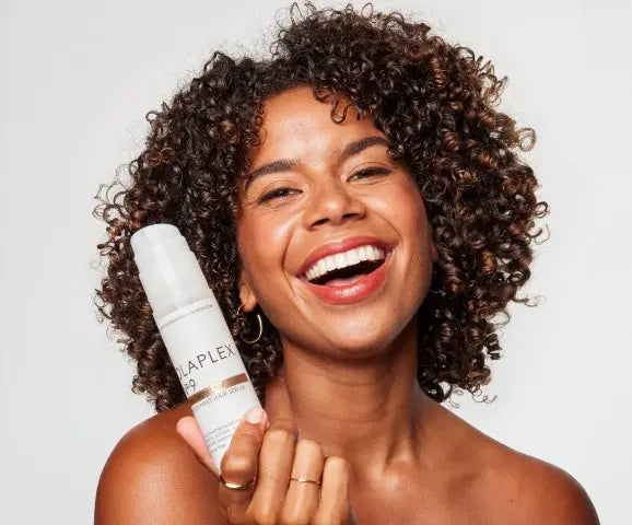 Smiling person with curly hair holds a white bottle labeled APPLE for damaged hair care.