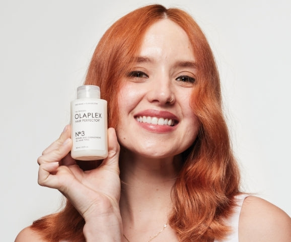 Smiling person with red hair holding Olaplex No. 3 hair perfector treatment bottle.