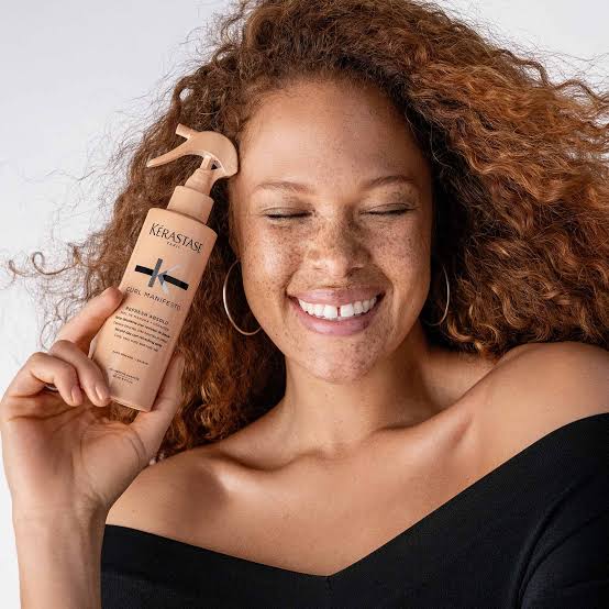 Smiling woman with curly hair holding Kerastase Curl Manifesto hair product for luxury curl care