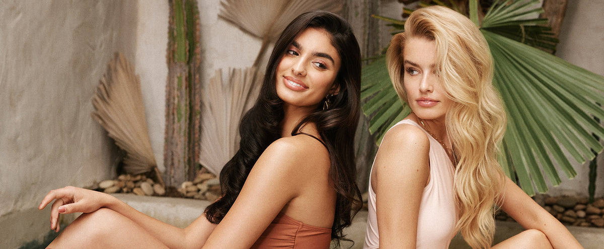 Moroccanoil features two women seated back-to-back in a tropical setting with lush greenery. One has dark hair, the other blonde, exuding a serene, natural elegance.