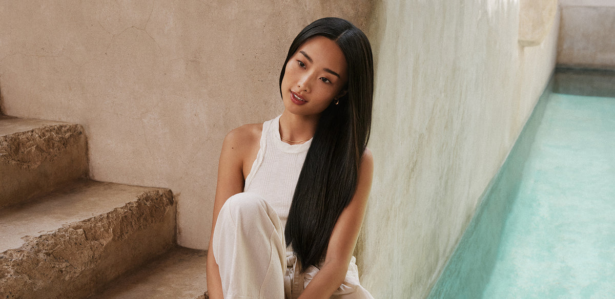 A serene scene of a woman with long, smooth, dark hair sitting on stone steps near a turquoise pool, representing the sleek, frizz-free results achieved with Moroccanoil Frizz Control.