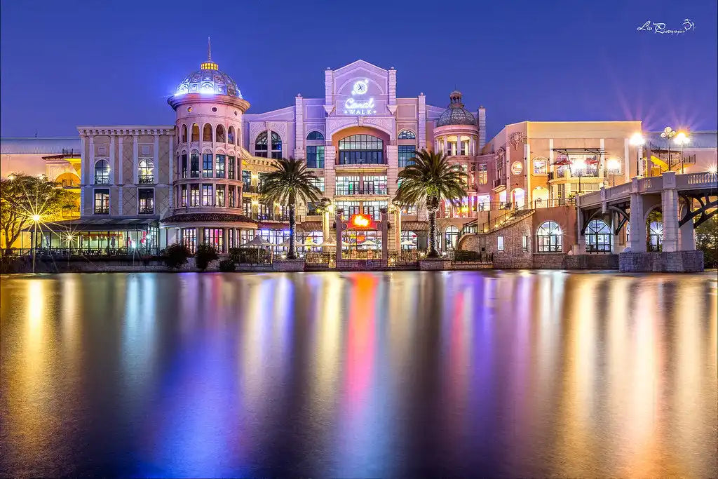 Illuminated Mediterranean-style waterfront shopping complex with ornate architecture and palm trees reflected in calm waters.