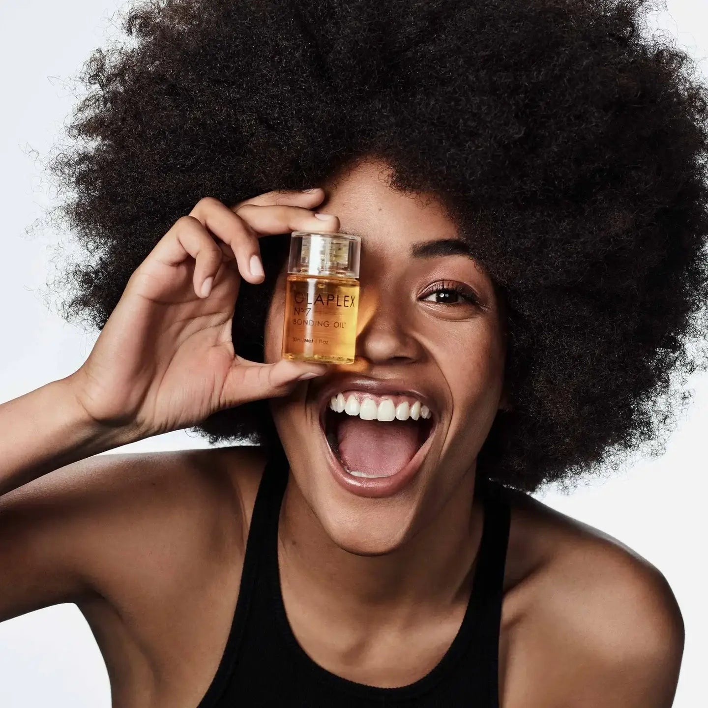 Smiling person with voluminous curly hair holding a small bottle of liquid in front of one eye.