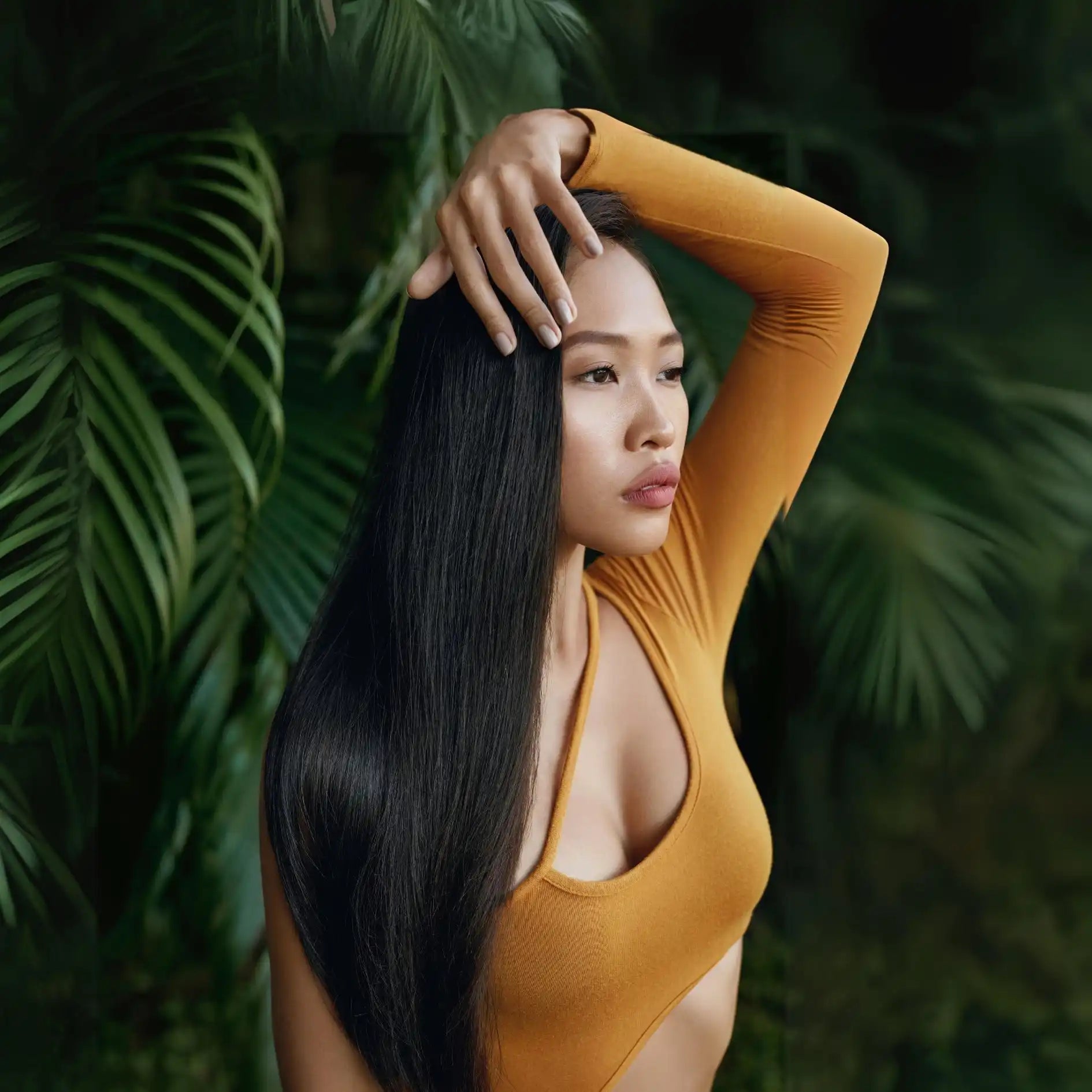 Woman with long black hair wearing a yellow top against a backdrop of green foliage.