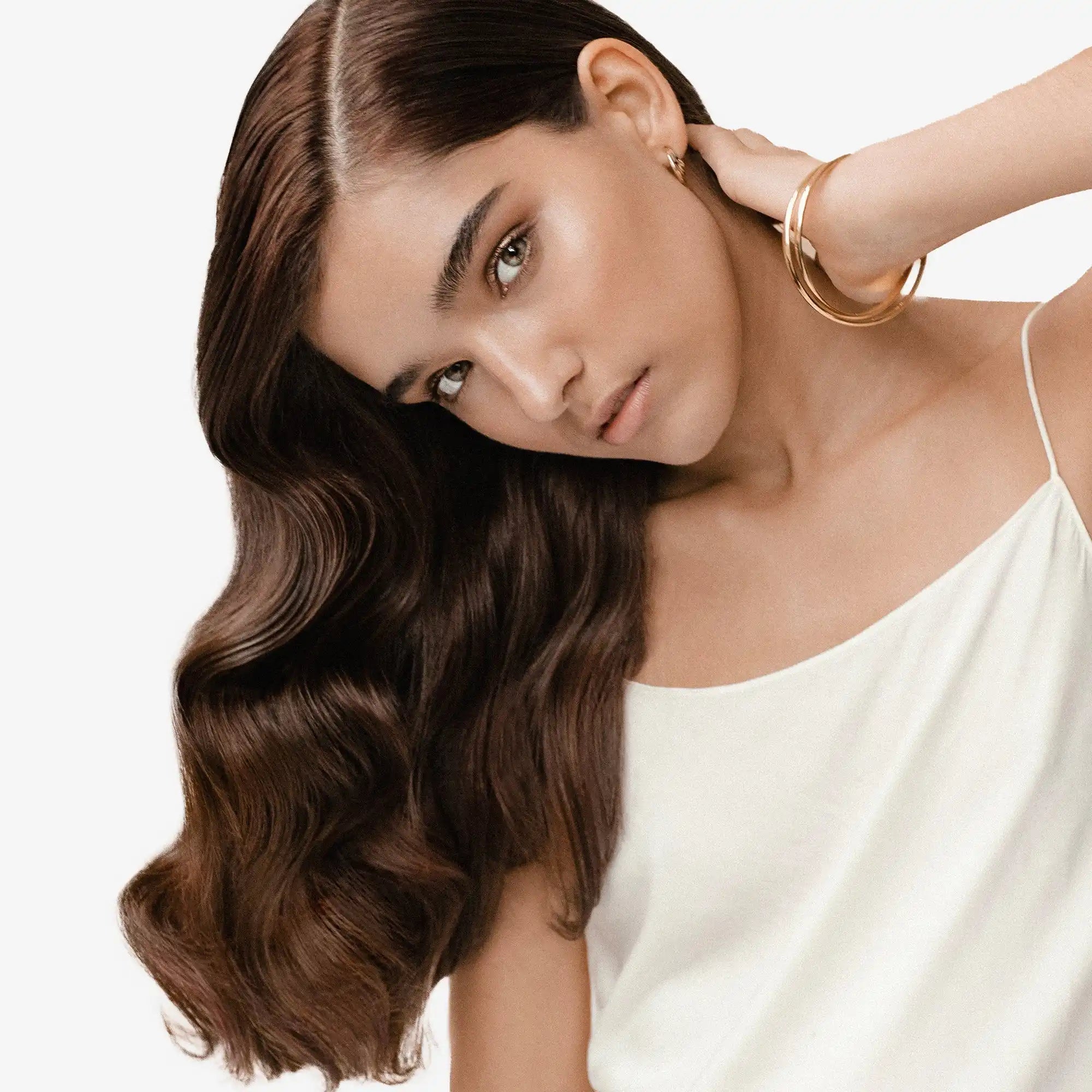 Woman with long, wavy brown hair and striking features wearing a white top and gold bracelet.